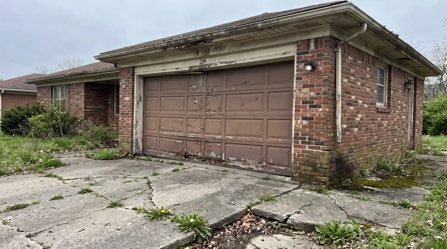 Before: A dented faded old brown garage door with peeling paint on a brick home with a cracked driveway, flat gray light